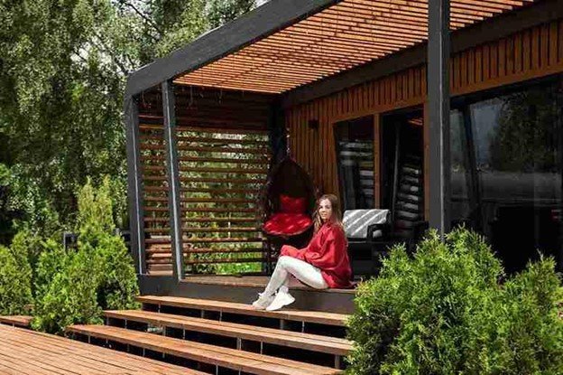Woman relaxing on a modern wooden deck with a pergola structure surrounded by greenery.